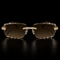 Rimless square sunglasses with gold-tone metal frames, crystal-studded brow bars, and brown gradient lenses, displayed on a reflective black surface.