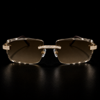 Rimless square sunglasses with gold-tone metal frames, crystal-studded brow bars, and brown gradient lenses, displayed on a reflective black surface.